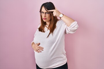 Pregnant woman standing over pink background pointing unhappy to pimple on forehead, ugly infection...