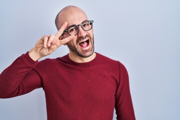 Young bald man with beard standing over white background wearing glasses doing peace symbol with fingers over face, smiling cheerful showing victory