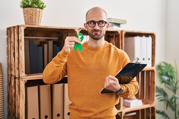 Young bald man with beard holding support green ribbon smiling looking to the side and staring away thinking.
