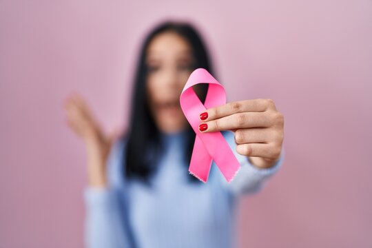 Hispanic woman holding pink cancer ribbon celebrating victory with happy smile and winner expression with raised hands