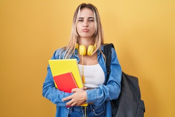 Young blonde woman wearing student backpack and holding books puffing cheeks with funny face. mouth inflated with air, crazy expression.