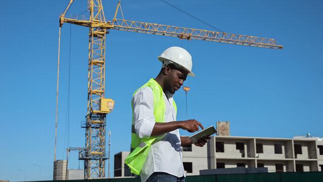 African American constructor monitors constructing process against blue sky making report on tablet. Black builder looks around entering data into device