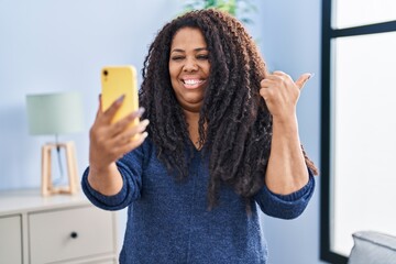 Plus size hispanic woman doing video call with smartphone pointing thumb up to the side smiling happy with open mouth