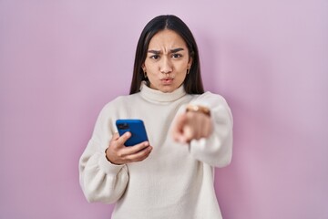 Young south asian woman using smartphone pointing with finger to the camera and to you, confident gesture looking serious