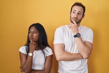 Interracial couple standing over yellow background thinking worried about a question, concerned and nervous with hand on chin