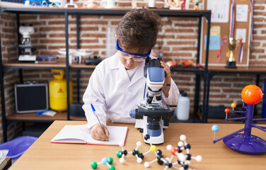 Adorable hispanic boy student using microscope writing on notebook at laboratory classroom