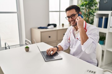 Young hispanic man wearing doctor uniform using laptop talking on the smartphone at clinic