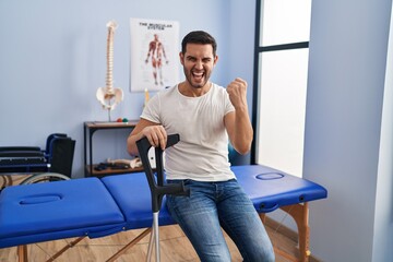 Young hispanic man with beard wearing crutches at rehabilitation clinic annoyed and frustrated shouting with anger, yelling crazy with anger and hand raised