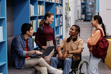 Diverse group of students with young man in wheelchair chatting cheerfully in college library,...