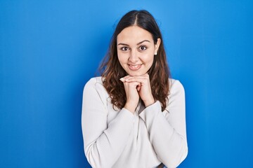 Young hispanic woman standing over blue background laughing nervous and excited with hands on chin looking to the side