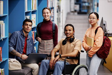 Diverse group of students with young man in wheelchair looking at camera in college library,...
