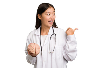 Young asian doctor woman holding a brain isolated points with thumb finger away, laughing and carefree.