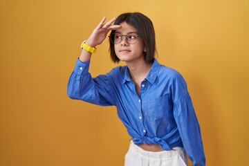 Young girl standing over yellow background very happy and smiling looking far away with hand over head. searching concept.