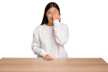 Young asian woman sitting on a table isolated having fun covering half of face with palm.