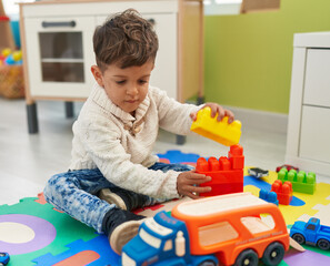 Adorable hispanic toddler playing with construction blocks sitting on floor at kindergarten