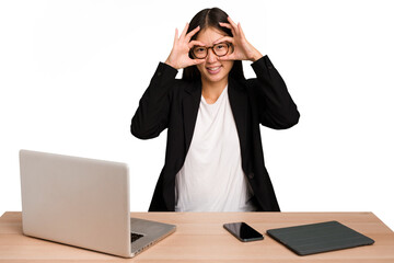 Young business asian woman sitting on a table isolated showing okay sign over eyes