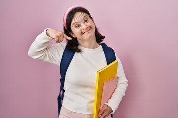 Woman with down syndrome wearing student backpack and holding books smiling cheerful showing and pointing with fingers teeth and mouth. dental health concept.