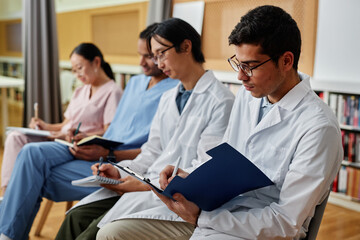 Group of medics in row taking notes during lecture or seminar, focus on young man holding clipboard...