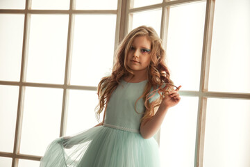 Portrait pensive little girl in art azure dress posing near large light window in room, looking at camera. Studio shot positive thoughtful kid lady. Child emotion concept. Copy text space for ad