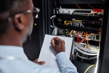 Close up of black man working with server cabinet in data center and taking notes on clipboard,...