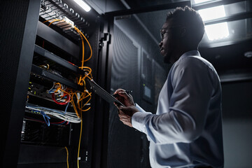Backlit side view portrait of black man working with server cabinet in data center and taking notes on clipboard, copy space - obrazy, fototapety, plakaty