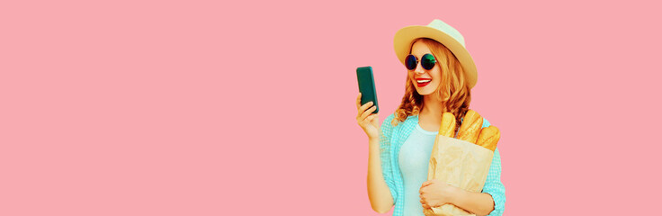 Portrait of happy smiling woman with smartphone holding grocery shopping paper bag with long white bread baguette on pink background, blank copy space for advertising text