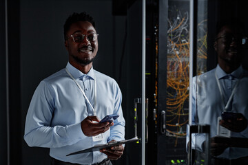 Waist up portrait of smiling black man as network engineer standing by server cabinet in data...