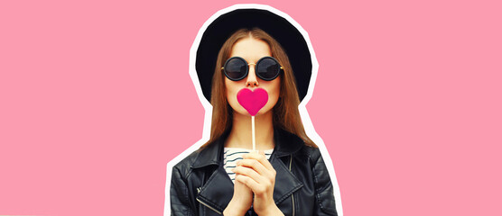 Portrait of stylish young woman with heart shaped lollipop posing wearing black round hat on pink...