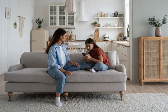 Sad teen girl holding mobile phone, experiencing cyberbullying on social media, sitting with mother on sofa at home. Mom helping daughter to cope with unrequited love. Teenage problems concept