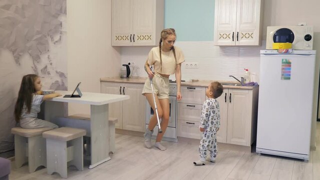 A young beautiful Caucasian girl moves around the kitchen of the house on one crutch, and dances with her little son while her daughter watches the tablet.Happy family. Leg injury, bandage.