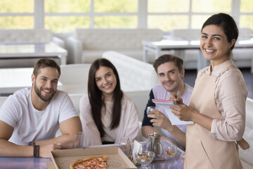 Portrait of pretty Indian waitress in apron serving group of happy young customers, guests of diner, cafe, sitting at table with pizza, hot drink, looking at camera, smiling
