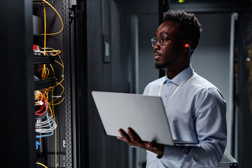 Waist up portrait of black man as network engineer in server room using laptop, minimal