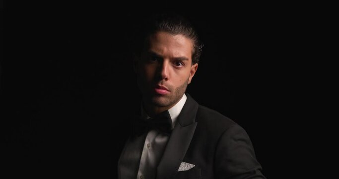 project video of sexy Lebanese man adjusting elegant black tuxedo and bowtie, making a hand gesture and inviting before walking away in darkness