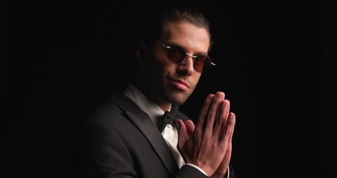 side view of attractive young groom in black tuxedo with sunglasses looking to side and rubbing palms in front of black background in studio