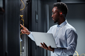 Side view portrait of young black man as network engineer working with servers in data center and...
