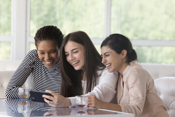 Three cheerful diverse girls making group video call on smartphone, using mobile phone in cafe, sitting at table in coffee shop, drinking tea, enjoying wireless communication, taking selfie