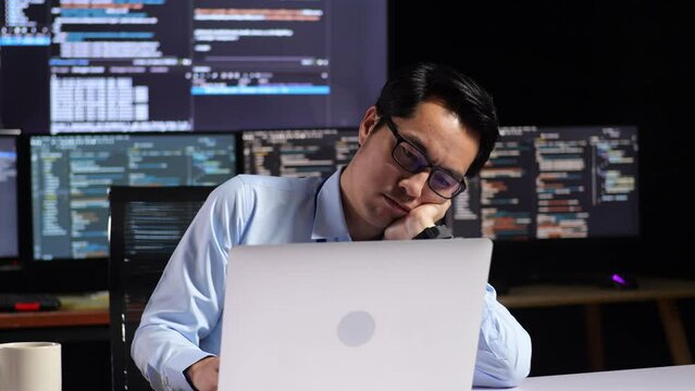 Asian Male Programmer Being Bored Writing Code By A Laptop Using Multiple Monitors Showing Database On Terminal Window Desktops In The Office
