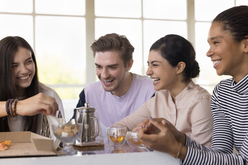 Diverse group of cheerful excited young friends gathering at table in cafe, talking, chatting, laughing, enjoying funny conversation, friendship, celebrating team success, drinking tea