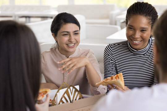 Happy young Indian and African female friends eating pizza in cafe, sitting at table, talking to friends, laughing, smiling, enjoying informal meeting, lunch break, friendship