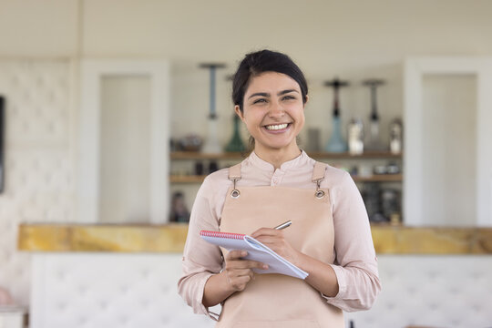 Happy beautiful Indian waitress in uniform apron holding pen, notebook for writing notes, looking at camera with toothy smile, enjoying job. Hookah lounge employee, barista professional portrait