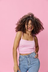 Woman with curly afro hair dancing in pink top and jeans on pink background, smile, copy place