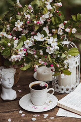 A beautiful postcard. A white coffee cup with a saucer, a statuette, candles, a book and a vase with a bouquet of blooming apple trees. Beautiful still life. Spring time. The concept of "Good morning"