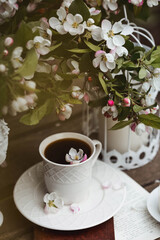 A beautiful postcard. A white coffee cup with a saucer, a statuette, candles, a book and a vase with a bouquet of blooming apple trees. Beautiful still life. Spring time. The concept of 