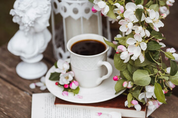 A beautiful postcard. A white coffee cup with a saucer, a statuette, candles, a book and a vase with a bouquet of blooming apple trees. Beautiful still life. Spring time. The concept of 