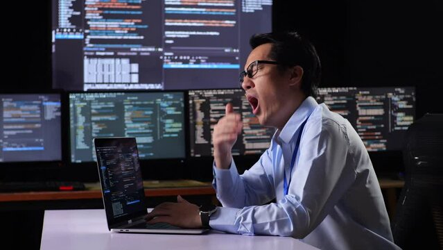 Side View Of Asian Male Programmer Yawning And Sleeping While Writing Code By A Laptop Using Multiple Monitors Showing Database On Terminal Window Desktops In The Office
