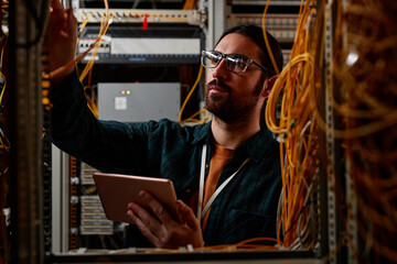 Front view portrait of bearded man setting up server while working with data network and holding tablet