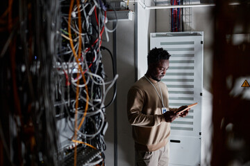 Moody wide angle shot of black man as system administrator standing in server room and holding tablet, copy space