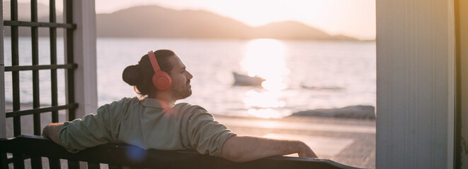 Portrait of a young man in bright large headphones listening to music, audiobook by the sea at sunset.
