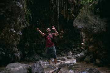 Naklejka na ściany i meble A young male tourist walks along a shallow mountain tropical river, shoots an action camera among the rocks.