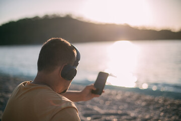 Portrait of a young man sitting on the seashore wearing headphones with a phone in his hands at...